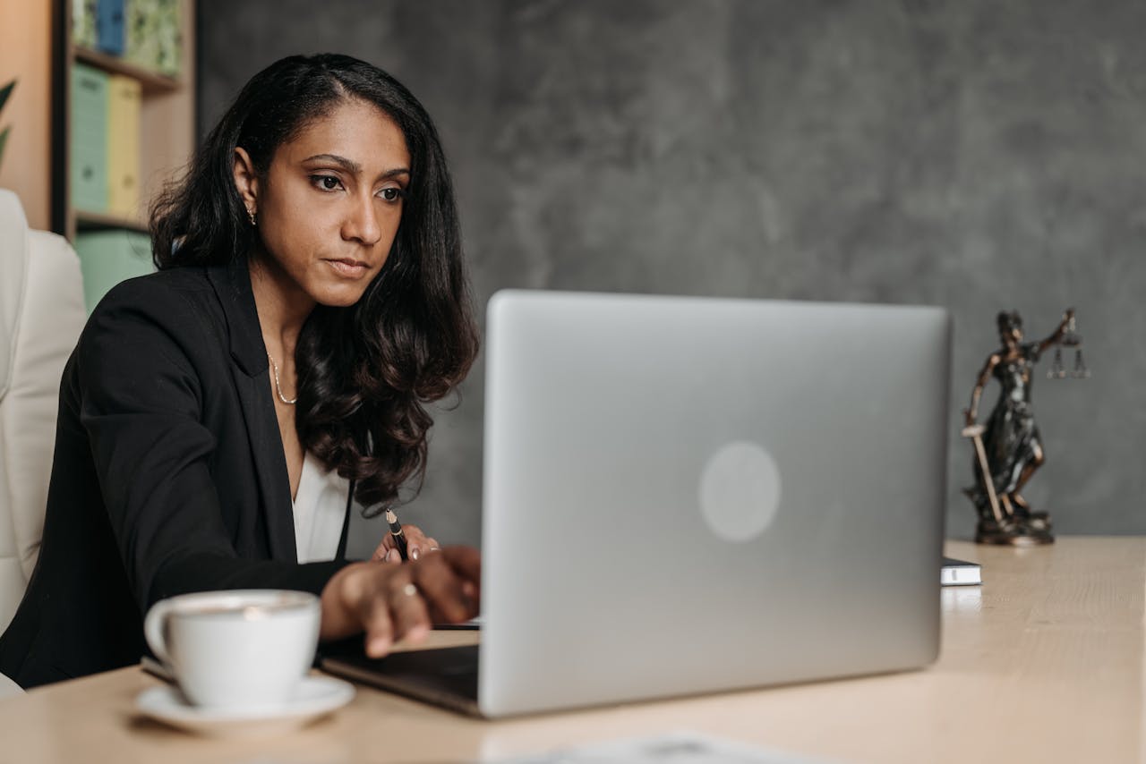 Professional woman in business attire using a laptop in a modern office setting with a cup of coffee and the scales of justice statue.